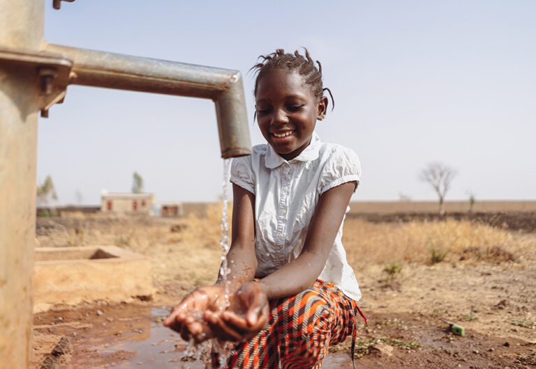 Gorgeous young black African village girl smiling happily at the clean water flowing out of the faucet; water scarcity concept